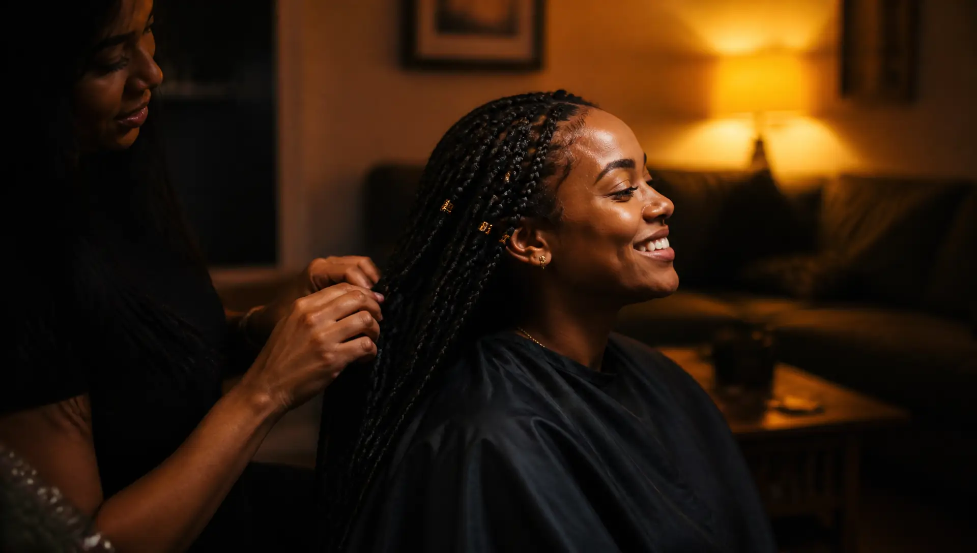 Braider's hands finishing long braids with gold cuffs in a warm living room